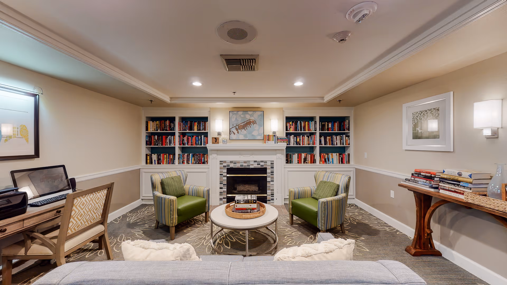 A cozy living room area in Island House Assisted Living featuring a fireplace with built-in bookshelves on either side filled with books. Two striped armchairs with green cushions face a round coffee table with books on it. A desk with a chair and printer is on the left side, and a wooden console table with more books and decorative items is on the right. The walls are painted beige with white trim, and framed artwork hangs on the walls. The room is softly lit with wall sconces and recessed ceiling lights.