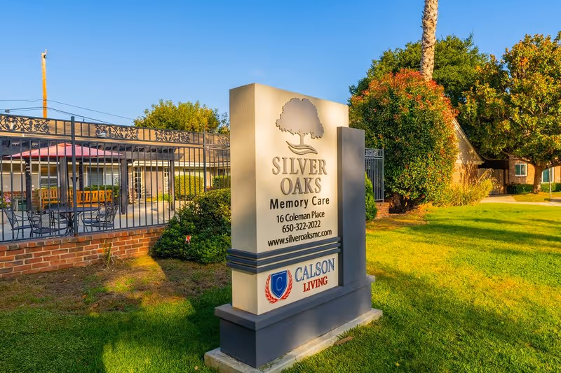 Outdoor view of the Silver Oaks Memory Care facility sign with contact information and logo, surrounded by green grass, trees, and a fenced patio area with tables and chairs in the background under a clear blue sky.