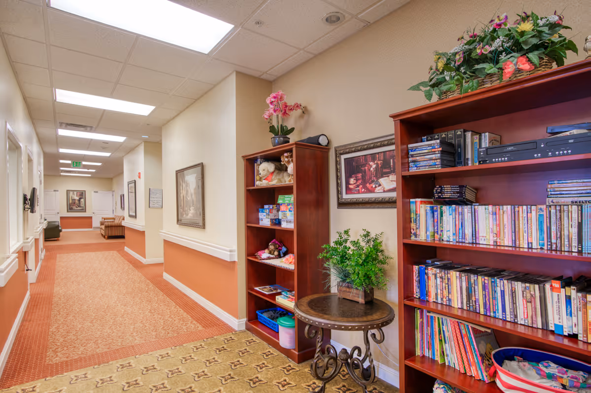 Carpeted interior hallway in a senior living facility with bookshelves full of books and games, a small table with a plant, framed art, and seating visible down the corridor.