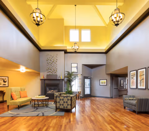 A spacious and well-lit living room area with high vaulted ceilings and three hanging chandeliers. The room features a fireplace with decorative art above it, a patterned armchair, a green loveseat with orange pillows, a round coffee table on a rug, and wooden flooring. There are framed pictures on the walls and a glass door leading outside.