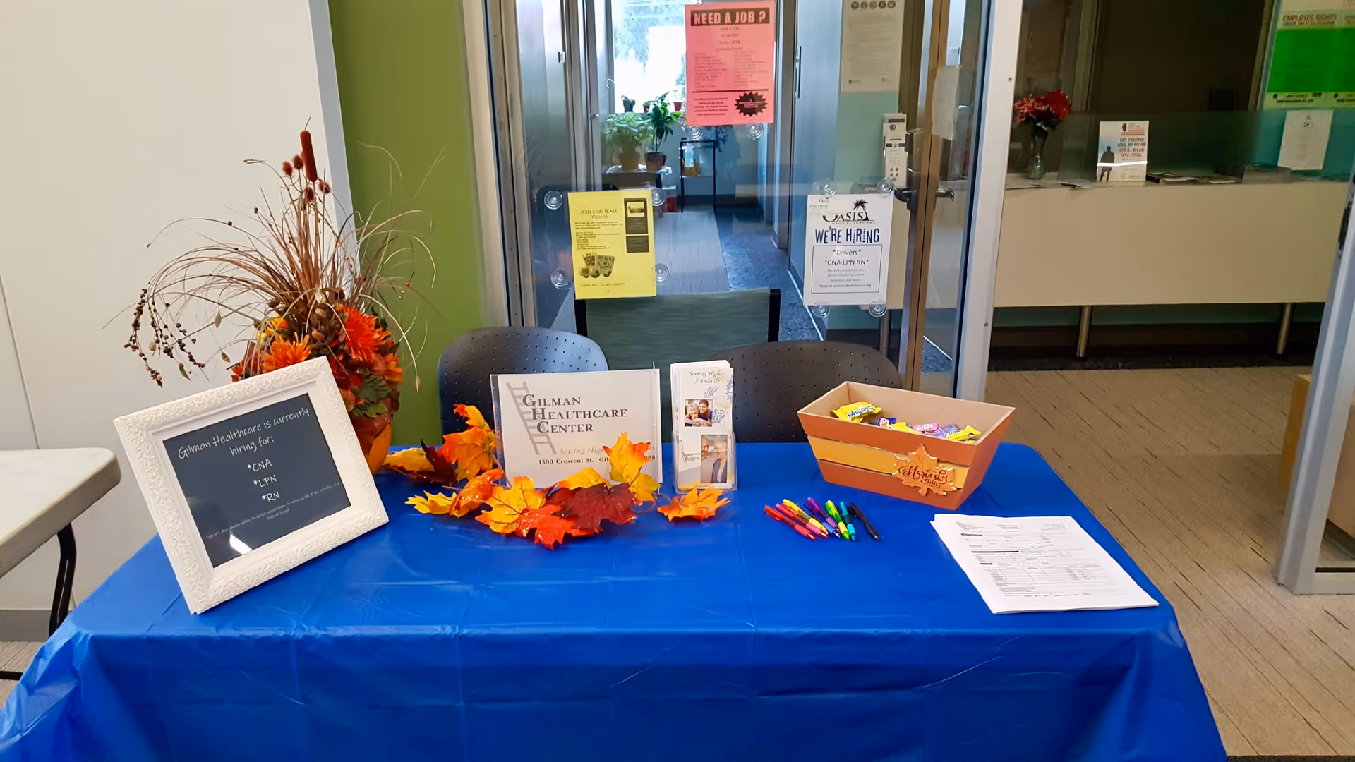 A blue tablecloth-covered table set up in a hallway or lobby area with informational brochures, a framed sign about Gilman Healthcare Center hiring for CNA, LPN, and RN positions, colorful pens, a box of candy, and autumn-themed decorations including artificial leaves and a floral arrangement. Behind the table are glass doors with hiring signs posted.