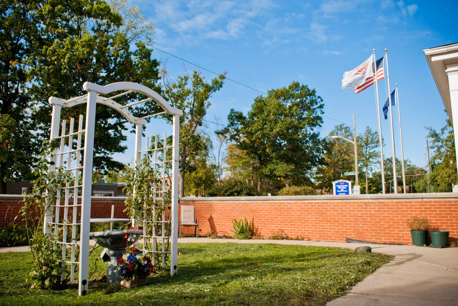 Outdoor garden area with a white wooden trellis adorned with climbing plants and a birdbath with flowers underneath. In the background, there is a red brick wall, a chair, three flagpoles with flags, and trees under a blue sky with some clouds.