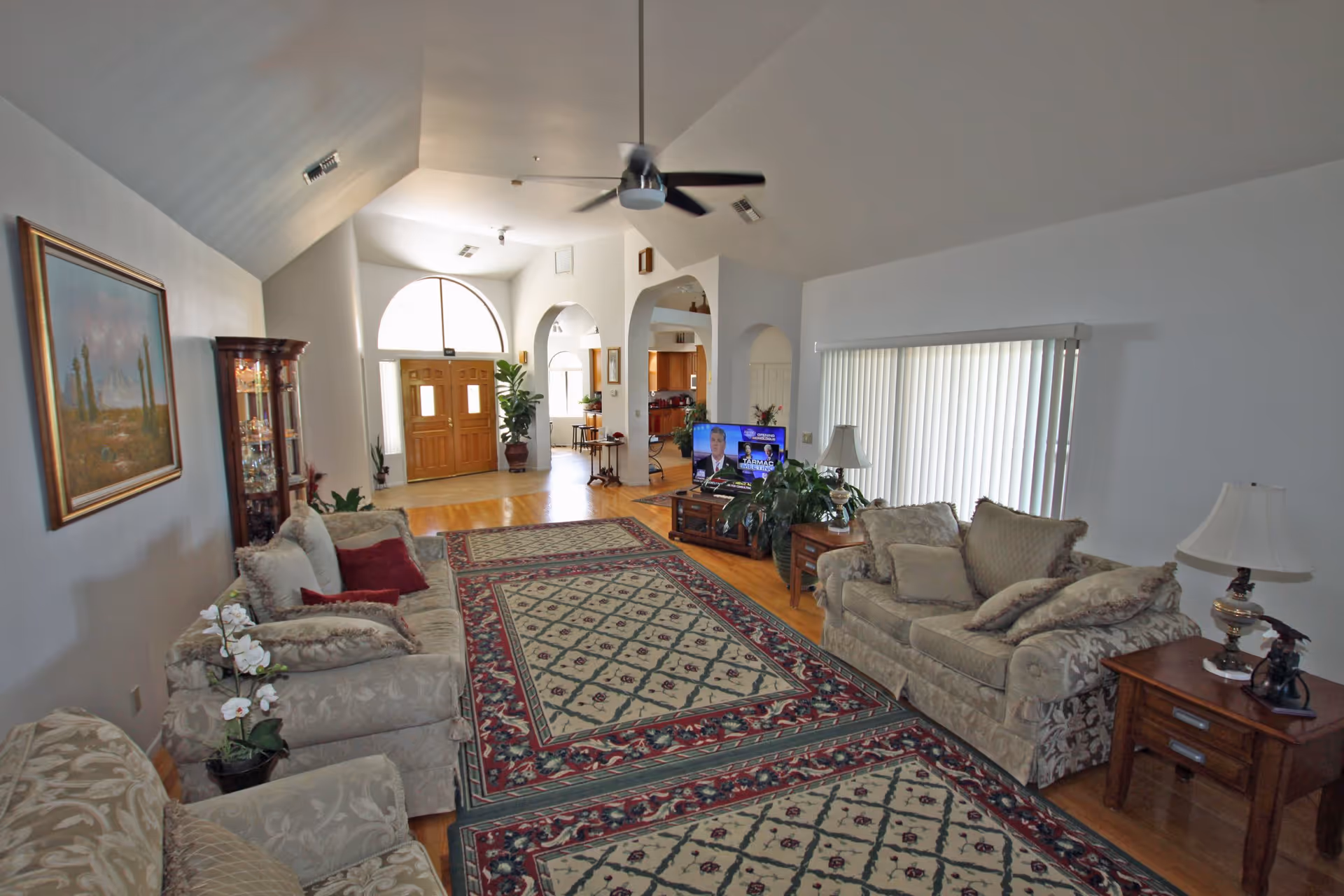 A spacious living room with high vaulted ceilings, featuring beige patterned sofas and armchairs arranged around a large area rug with a red and green floral pattern. There is a wooden coffee table with a TV on it, a side table with a lamp, and a large window with vertical blinds. The room has hardwood floors and white walls, with a painting on one wall and a glass display cabinet in the corner. The entrance door with an arched window above it is visible in the background along with an open archway leading to another room.