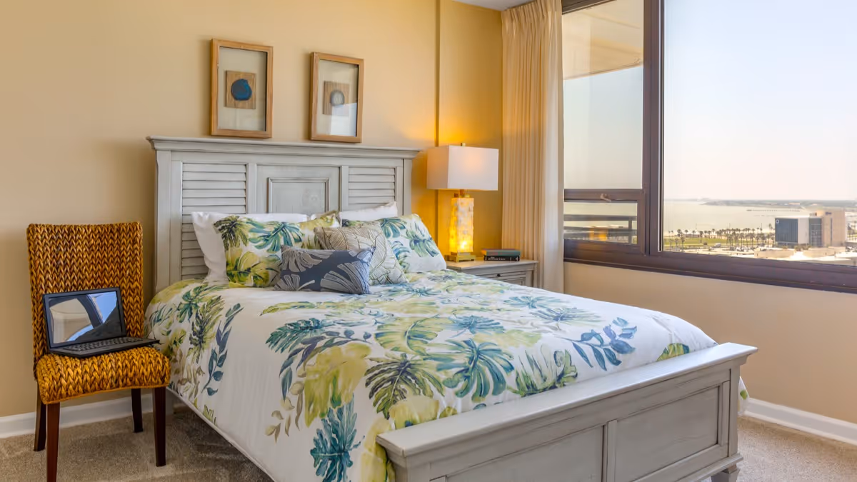 A bright bedroom with a large window showing a city and waterfront view. The room features a white wooden bed frame with a floral patterned bedspread in green and blue tones. There is a wicker chair with a laptop on it next to the bed, and a nightstand with a lamp and books. Two framed artworks hang above the bed.