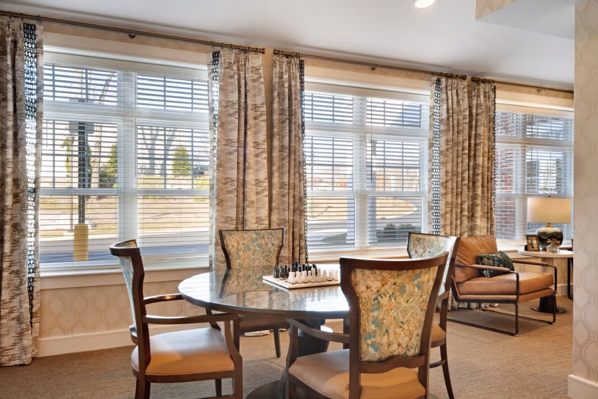 A cozy sitting area with a round table and four upholstered chairs featuring a floral pattern. A chess set is placed on the table. Behind the table are large windows with white blinds and patterned curtains. To the right, there is a comfortable armchair with a cushion and a side table with a lamp and framed photo. The room has beige carpet and wallpaper with a subtle geometric pattern.