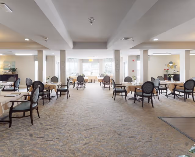 A spacious dining room with multiple tables covered in beige tablecloths, each surrounded by dark wooden chairs with cushioned seats and backs. The room is well-lit with natural light coming through large windows at the far end. The walls are light-colored, and there are decorative elements such as plants, paintings, and mirrors. The carpet has a subtle pattern, and the ceiling features recessed lighting and structural beams.