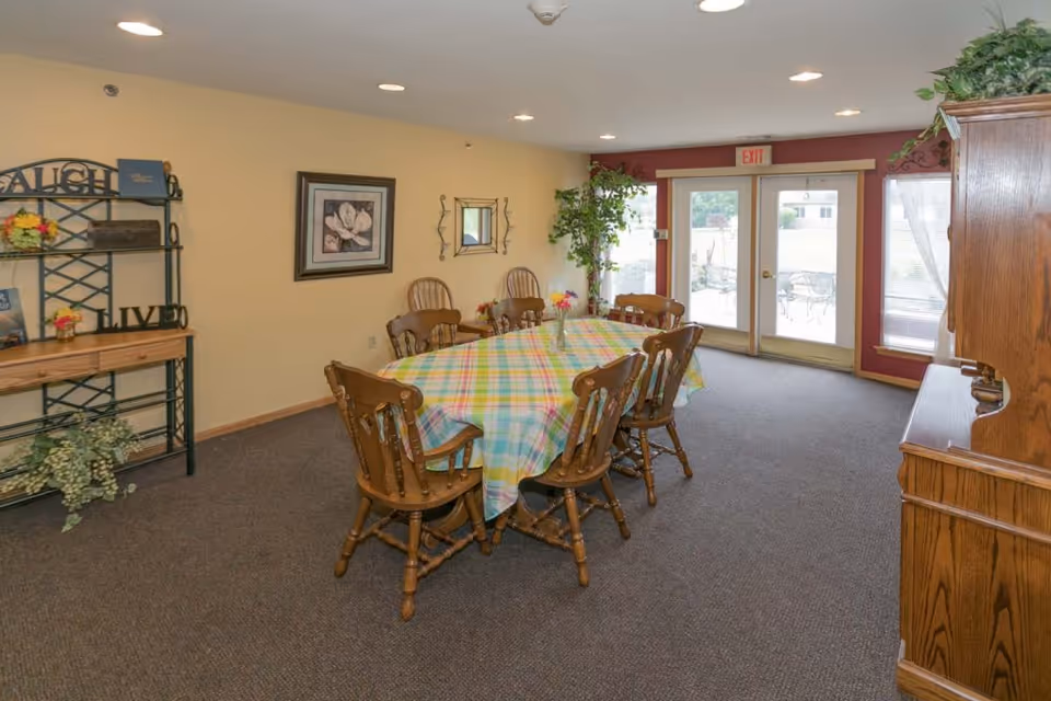 A dining room with a rectangular table covered with a colorful plaid tablecloth surrounded by eight wooden chairs. The room has beige walls with framed artwork and a mirror, a wooden shelving unit with decorative items, a wooden cabinet, and a large window and glass door leading outside. There is a potted plant near the door and carpeted flooring.