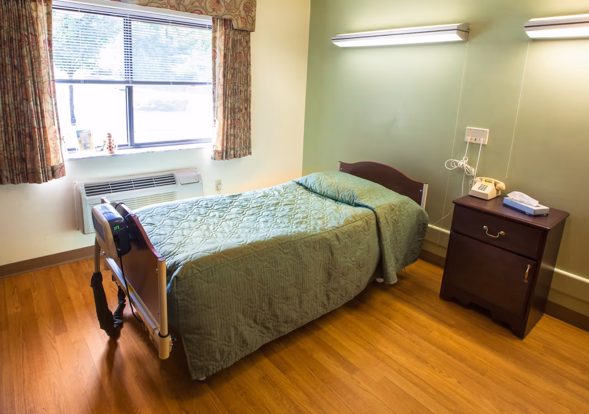 A simple bedroom with a single bed covered in a green quilted blanket and matching pillow. Next to the bed is a wooden nightstand with a beige rotary phone and a box of tissues. The room has wooden flooring, a window with floral curtains, and two wall-mounted lights above the bed.