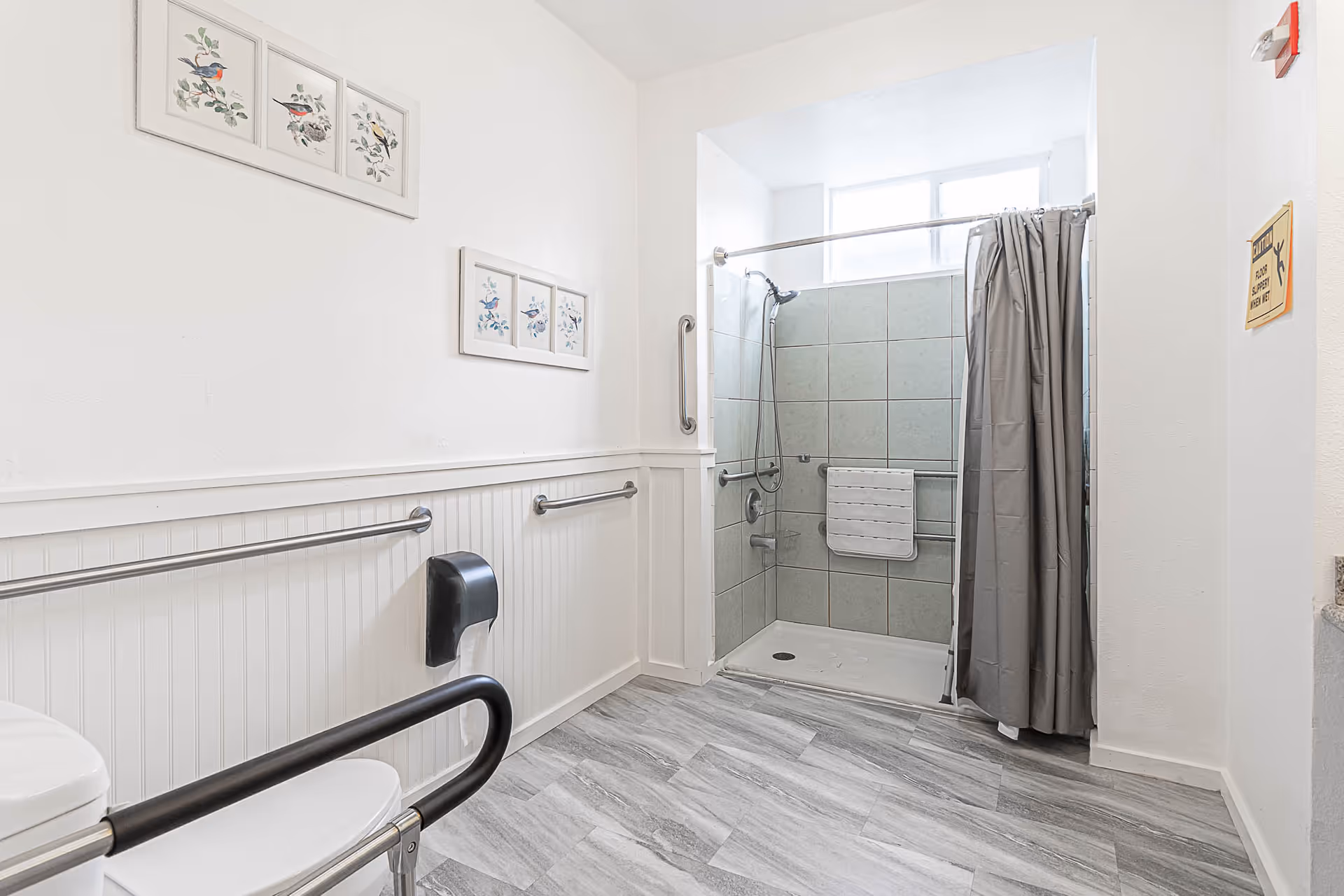 A clean, accessible bathroom with a walk-in tiled shower featuring a handheld showerhead and grab bars. The bathroom has light gray wood-look flooring, white walls with wainscoting, and framed bird artwork on the walls. There is a toilet with a black safety rail and a black toilet paper dispenser mounted on the wall.