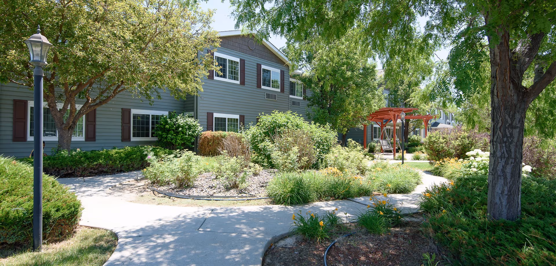 Landscaped courtyard with a winding concrete path, trees, shrubs, and a two-story residential building with a pergola in the background.