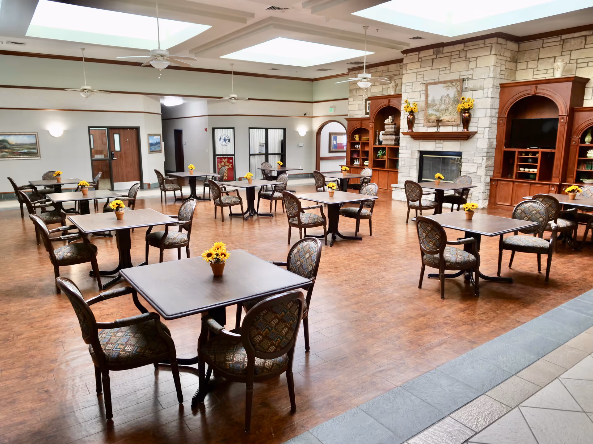 A spacious dining room with multiple square tables, each surrounded by four upholstered chairs. Each table has a small pot of yellow flowers. The room features a stone fireplace with wooden built-in shelves on either side, decorated with vases and a painting above the mantel. The floor is wooden, and the ceiling has skylights and ceiling fans.