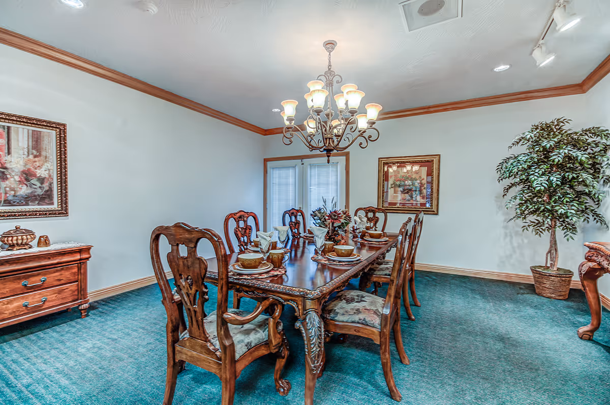 A formal dining room with a long wooden dining table set with plates, bowls, and napkins. The room features ornate wooden chairs with floral upholstery, a chandelier hanging from the ceiling, framed artwork on the walls, a wooden sideboard, and a potted plant in the corner. The carpet is green and the walls are light-colored with wooden trim.