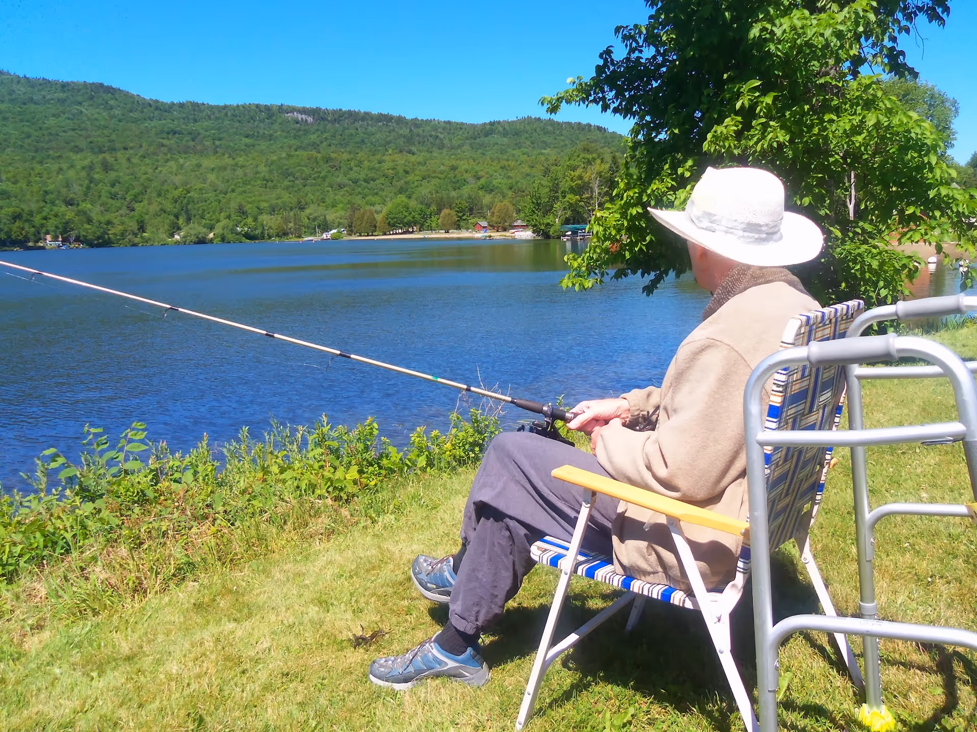 An elderly person wearing a white hat and beige jacket is sitting on a folding chair by a lake, fishing with a fishing rod. There is a walker next to the chair, and the background shows a calm lake with green trees and hills under a clear blue sky.