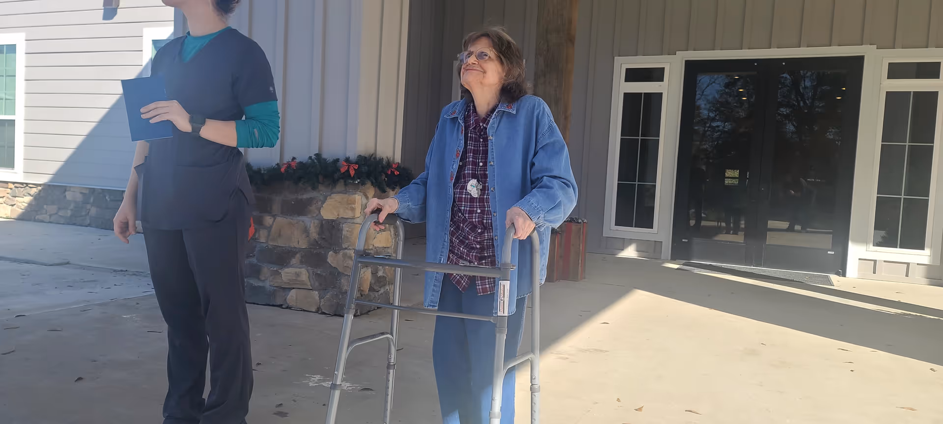 An elderly woman using a walker stands outside a building entrance, accompanied by a caregiver holding a notebook. The building has stone and siding exterior with glass double doors and windows.