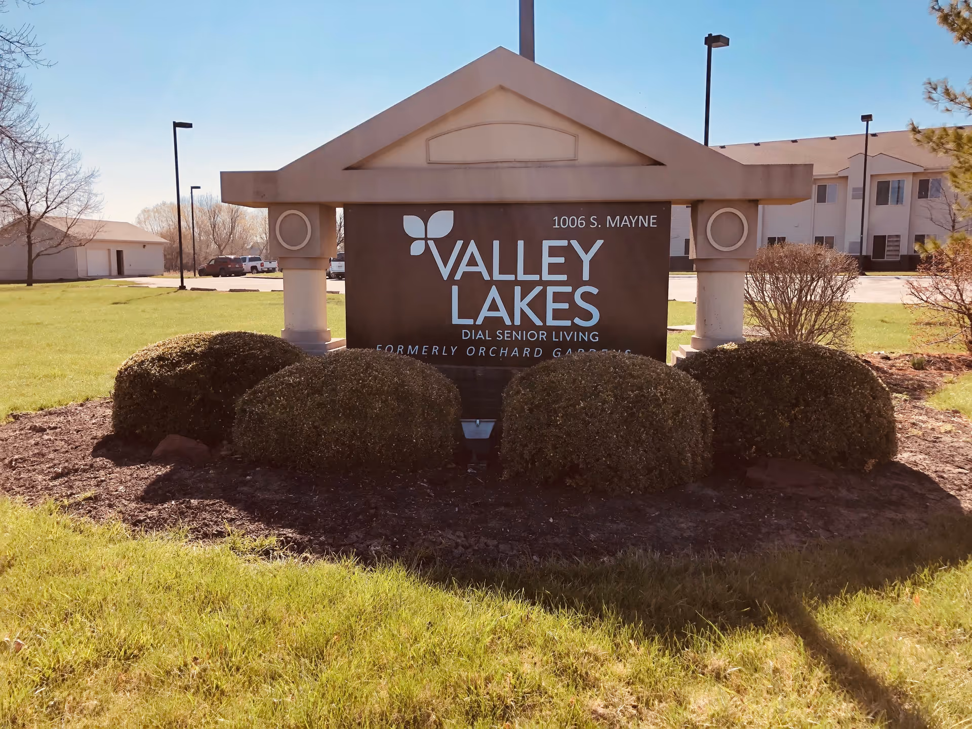 Outdoor view of the entrance sign for Valley Lakes Dial Senior Living, formerly Orchard Gardens, located at 1006 S. Mayne. The sign is surrounded by trimmed bushes and grass, with a building and parking lot visible in the background under a clear blue sky.