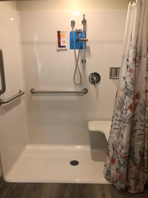 Accessible shower area with a handheld showerhead, grab bars on the walls, a fold-down seat, and a floral-patterned shower curtain partially drawn to the side.