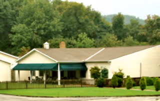 A single-story senior living facility with a beige exterior, a green awning over the entrance, and a manicured lawn with a low black fence.