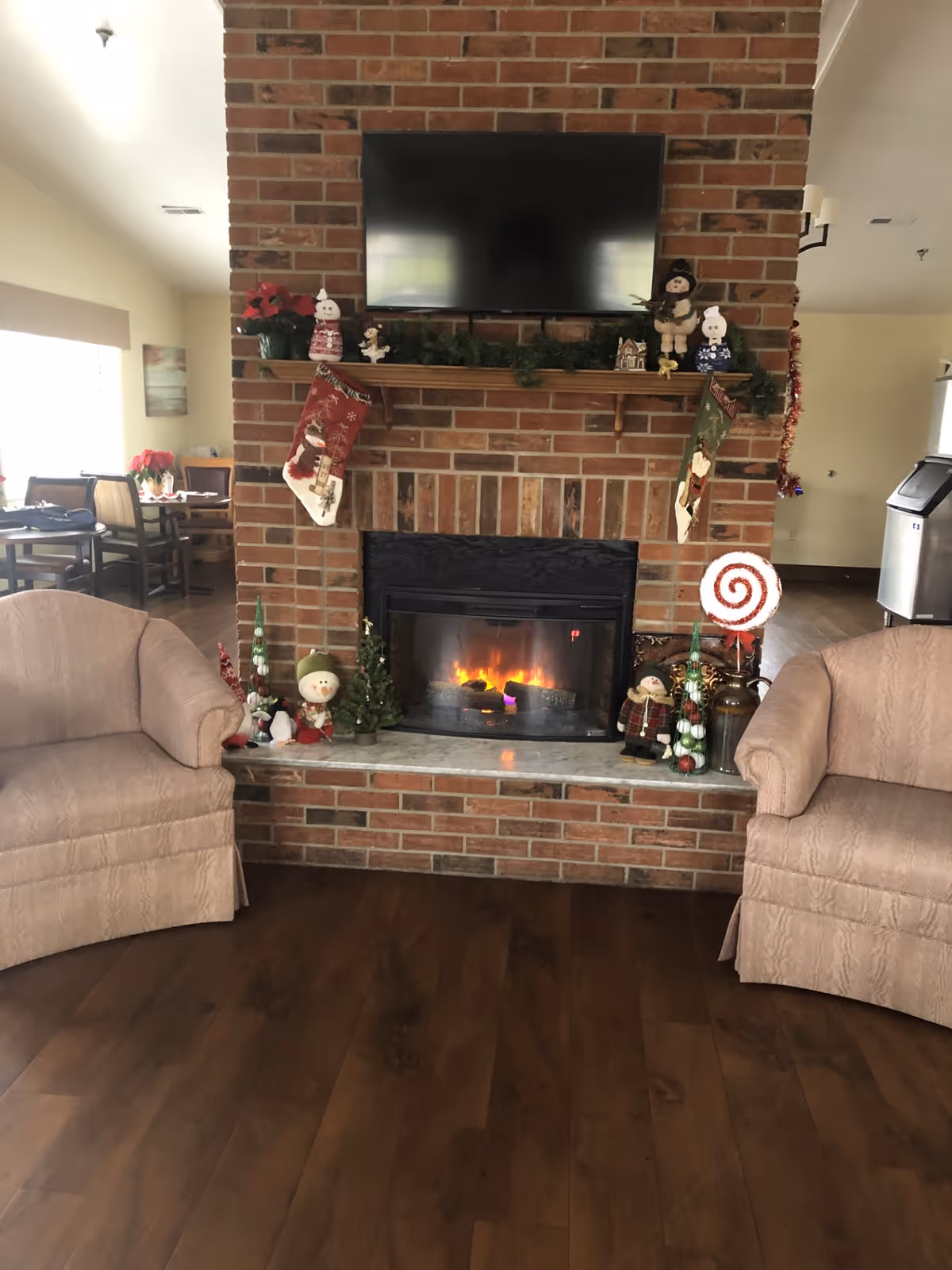 A cozy living room area with a brick fireplace in the center, decorated with Christmas stockings, small holiday figurines, and greenery. Above the fireplace is a mounted flat-screen TV. Two beige upholstered armchairs are positioned on either side of the fireplace. The room has wooden flooring and a dining area is visible in the background to the left.