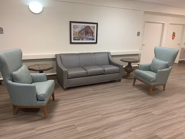 A seating area in a senior living facility with a gray three-seat sofa flanked by two light blue armchairs with cushions. There are two small round wooden side tables next to the sofa. The floor has wood-like vinyl planks, and the walls are light-colored with a framed picture of a cabin hanging above the sofa. Two doors are visible in the background, one with a small owl decoration.