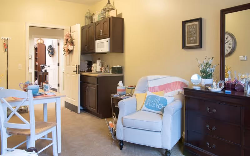 A cozy interior room featuring a white armchair with a blue 'hello' pillow and a pink and white throw blanket. To the right is a dark wooden dresser with decorative items and a mirror above it. On the left side, there is a small dining table with two white chairs and place settings. In the background, a kitchenette with dark wooden cabinets, a microwave, and a countertop is visible. The door is open, showing another room decorated with a wreath.