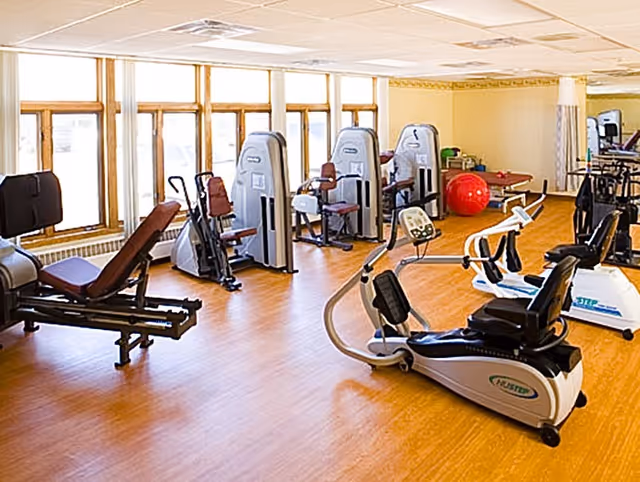A well-lit exercise room with various fitness equipment including recumbent bikes, weight machines, and a large red exercise ball. The room has large windows allowing natural light to fill the space and wooden flooring.