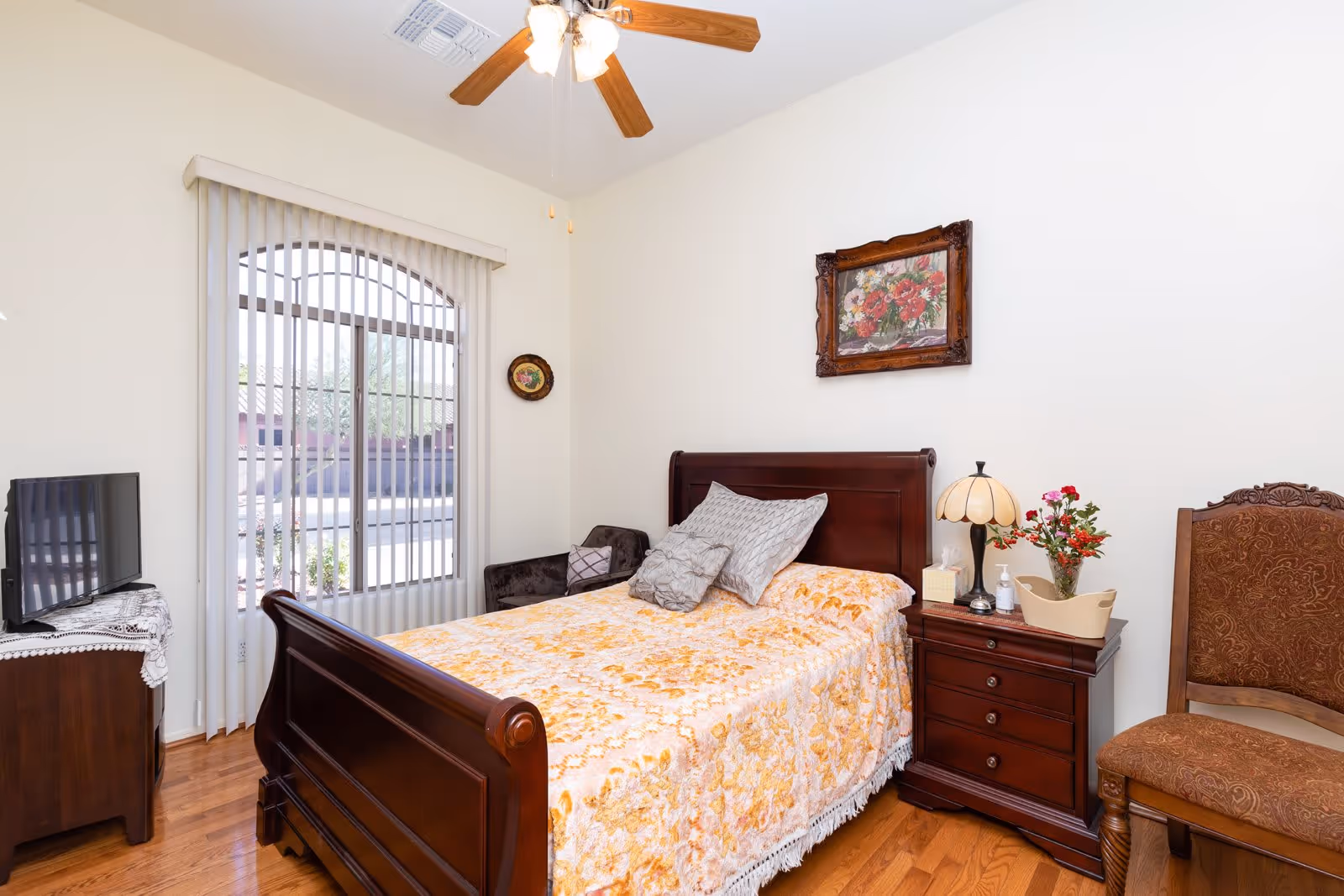 A tidy bedroom with a wooden bed covered by a floral bedspread, a nightstand with lamp and flowers, a chair, and a window with vertical blinds.