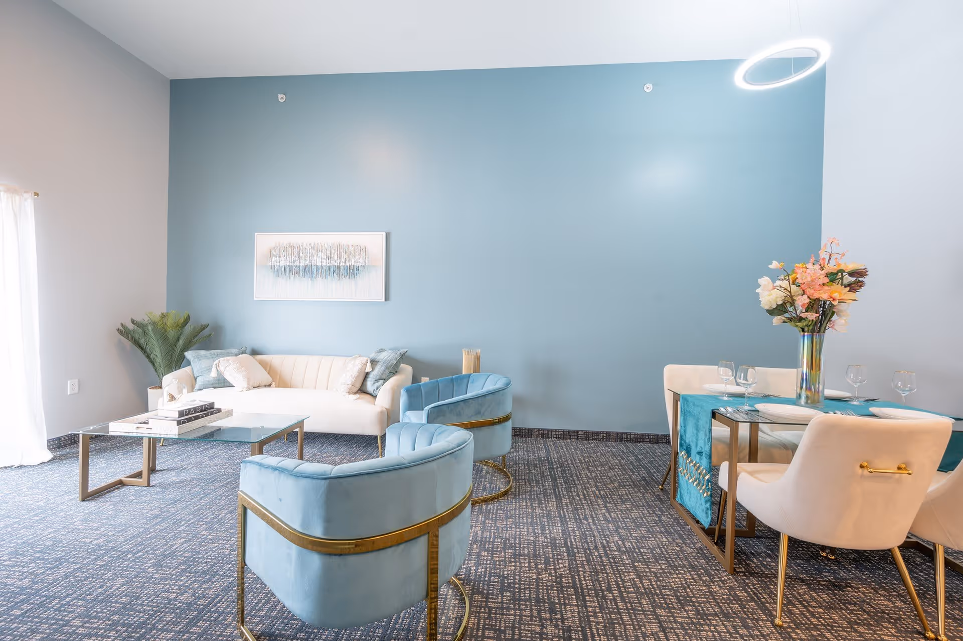 A modern living and dining area with a light blue accent wall. The living area features a white sofa with cushions, two light blue velvet chairs with gold accents, a glass coffee table with books, and a potted plant in the corner. The dining area has a glass table with a blue table runner, four cream-colored chairs, and a vase with flowers. The room has a patterned carpet and a circular ceiling light.