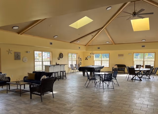 Spacious common area with high vaulted ceiling featuring skylights and ceiling fan. The room has multiple seating arrangements including wicker chairs with cushions, a pool table, and several tables with chairs. Large windows allow natural light to fill the room, and the walls are decorated with clocks and wall art.