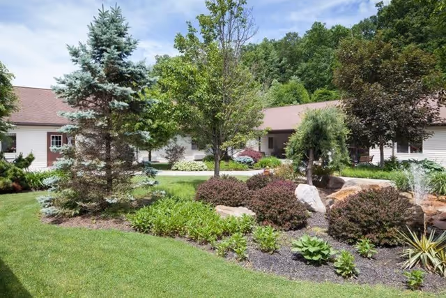 A landscaped garden area with various trees, bushes, and plants in front of a single-story building with a brown roof, under a partly cloudy sky.
