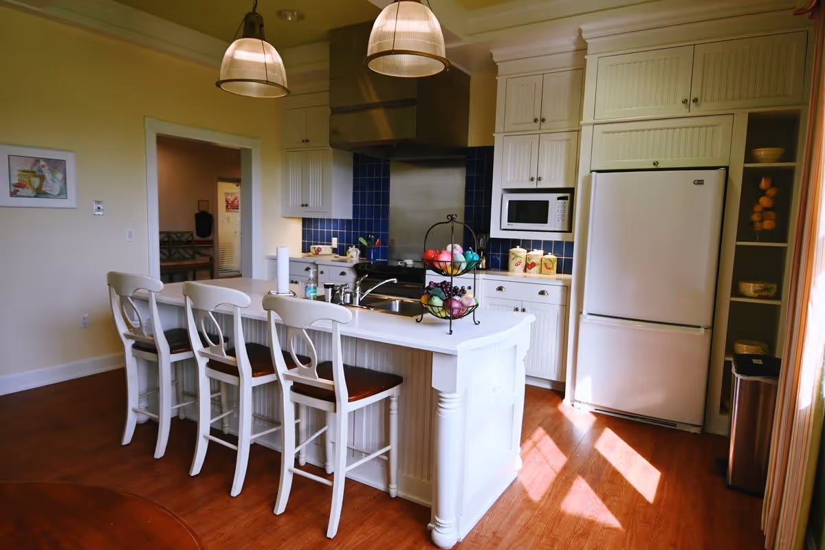 A bright kitchen with white cabinetry and a large island featuring four white wooden bar stools with dark seats. The kitchen has a stainless steel range hood, a white refrigerator, a microwave, and a blue tiled backsplash. Two pendant lights hang above the island, which has a sink and a two-tier fruit basket with colorful fruit. The floor is wooden, and sunlight streams in from the right side.