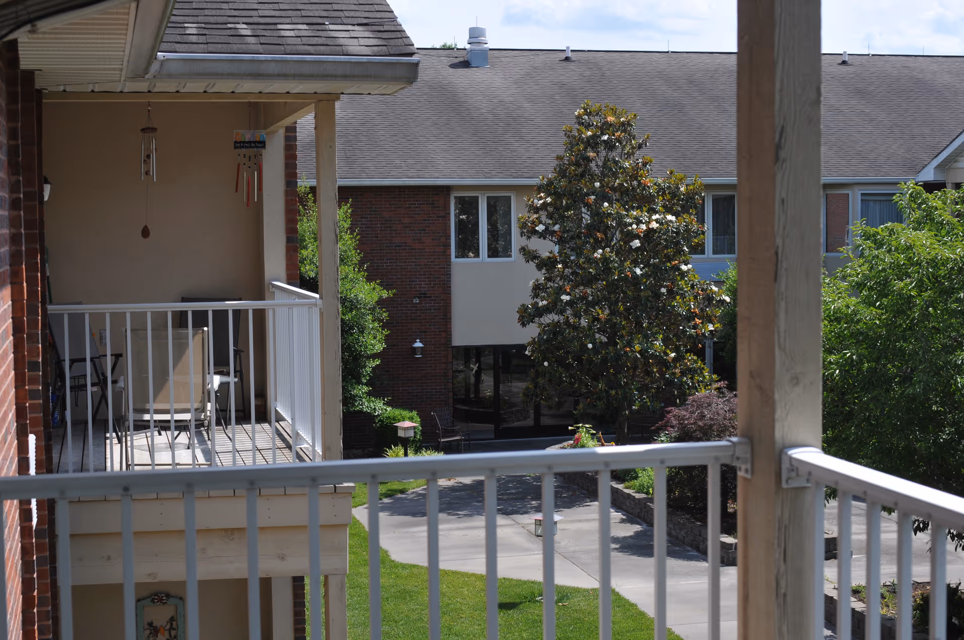 View from a balcony overlooking a courtyard area with a large flowering tree, green bushes, and a walkway. The balcony has white railings and outdoor chairs. The background shows a two-story brick building with windows and a sloped roof under a partly cloudy sky.
