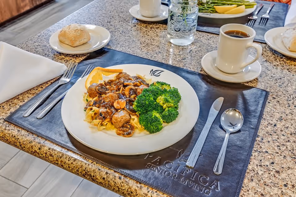 A dining table set with a plate of meatballs and mushrooms over noodles with broccoli on the side, a bread roll on a small plate, a cup of black coffee, and silverware on a dark placemat embossed with the text 'PACIFICA SENIOR LIVING'.