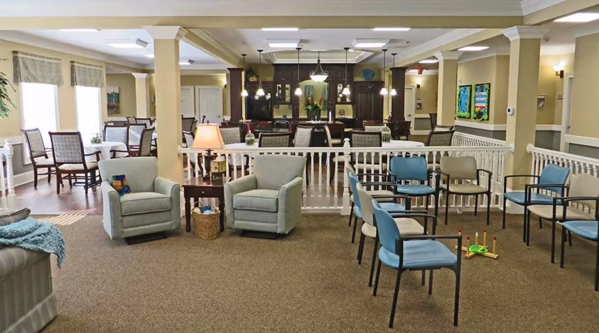 A spacious senior living common area with beige walls and carpeted floor. The room features multiple seating arrangements including light blue armchairs, blue and beige chairs arranged in a circle, and dining tables with cushioned chairs near large windows. There is a small table with a lamp and a basket between two armchairs. The background shows a kitchen area with dark wood cabinets and hanging pendant lights. The room is well-lit with ceiling lights and natural light from the windows.