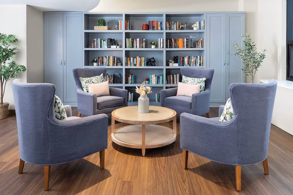 A cozy sitting area with four blue upholstered armchairs arranged around a round wooden coffee table with a vase of dried flowers. Behind the chairs is a large built-in bookshelf filled with books and decorative items. Two potted plants are placed on either side of the bookshelf, and the floor is wooden.