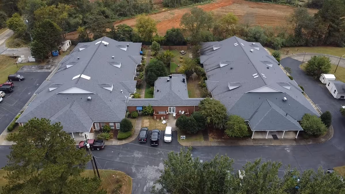 Aerial view of Highlands Senior Living Rome showing two large connected buildings with gray roofs, surrounded by trees and parking areas with several cars parked.