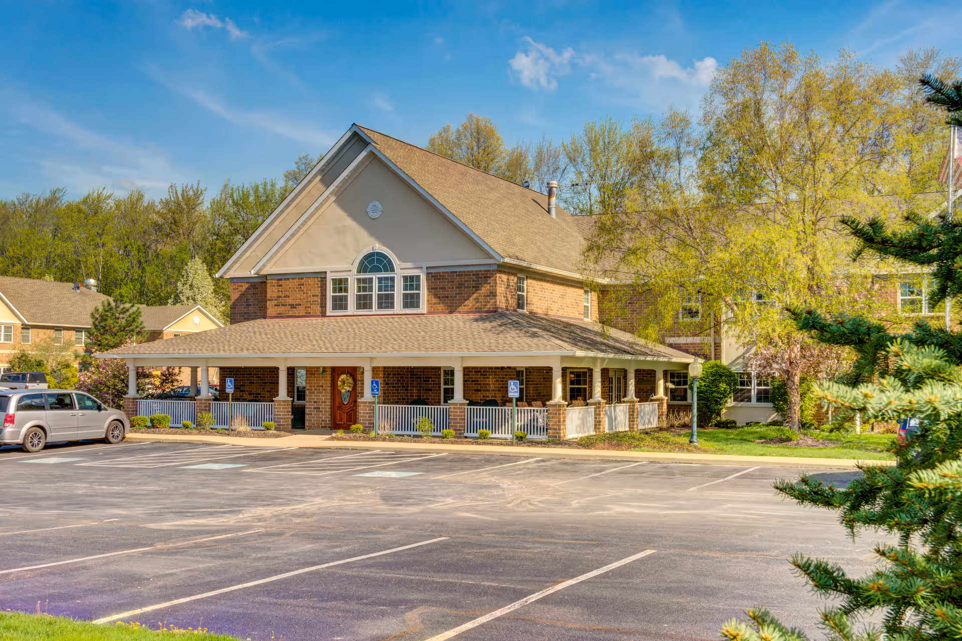 Exterior view of a two-story senior living facility building with a brick and beige facade, a covered porch with white railings, several handicap parking spaces in front, surrounded by trees and greenery under a blue sky.