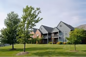 Exterior view of a senior living facility building with multiple units, featuring balconies and surrounded by a well-maintained lawn and several trees under a partly cloudy sky.
