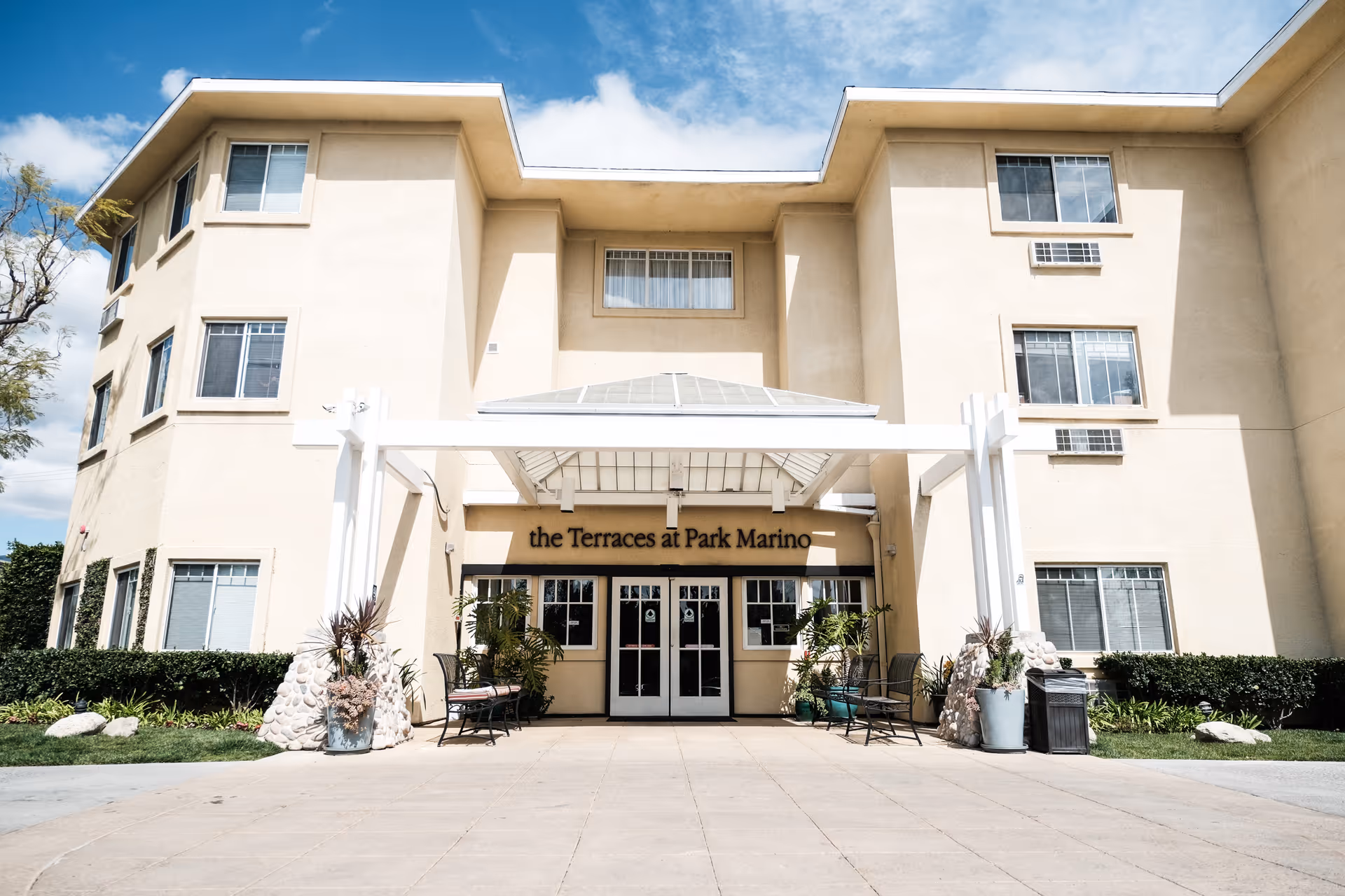 Front entrance of The Terraces at Park Marino building with a white pergola, glass double doors, and potted plants.