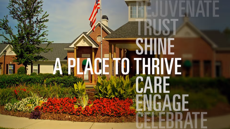 Exterior view of a senior living facility building with a well-maintained garden featuring red and white flowers and green shrubs. An American flag is flying on a flagpole near the entrance. The building has a brick facade with white trim and a covered entryway.