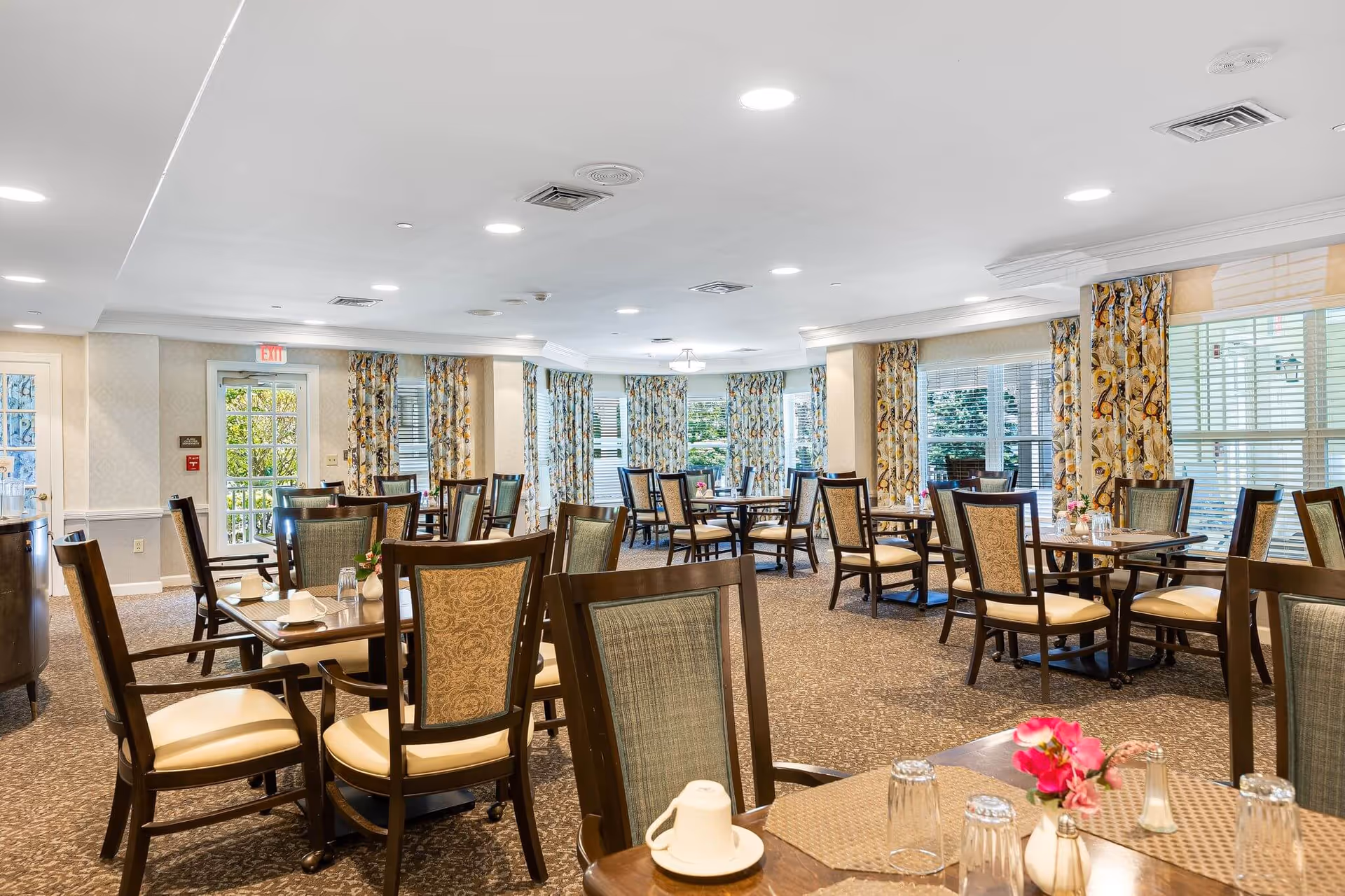 A bright and spacious dining room with multiple wooden tables and chairs arranged neatly. The room features large windows with floral curtains allowing natural light to fill the space. Tables are set with placemats, glasses, and small flower arrangements. The carpeted floor and soft lighting create a warm and inviting atmosphere.