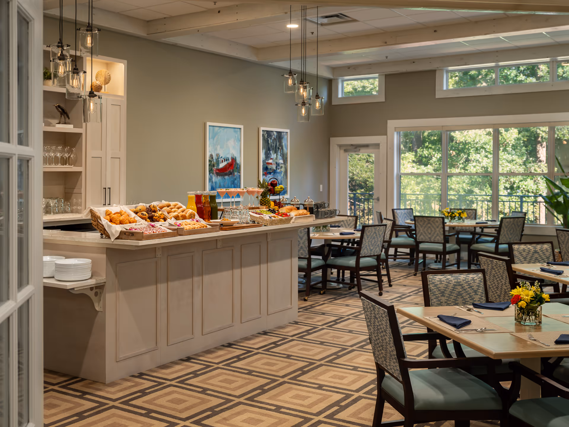 Bright dining room with a central buffet island displaying pastries, juices and dishes, surrounded by tables and chairs and large windows showing greenery outside.