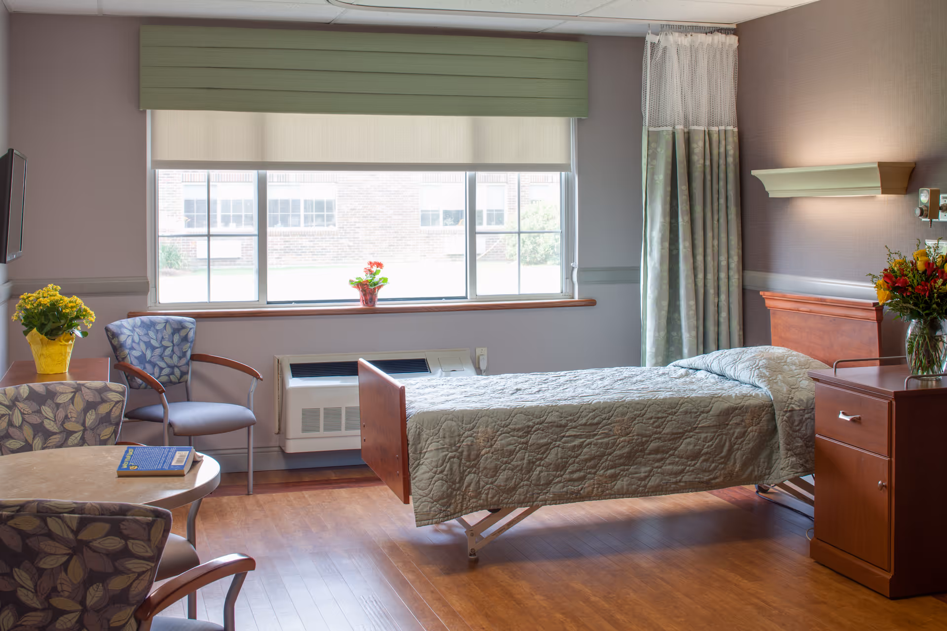 A bright and clean senior living bedroom with a single bed covered in a green quilt, a wooden nightstand with a vase of flowers, two floral patterned chairs around a small round table with a book on it, a large window with a green valance and a white roller shade, and a heating/cooling unit beneath the window.