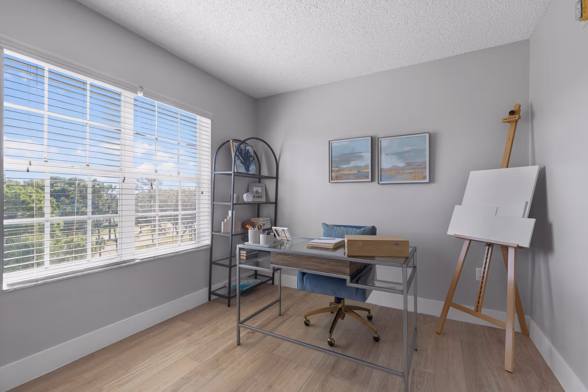 A bright room with a large window covered by white blinds, a modern glass and metal desk with a blue cushioned chair, a black metal shelving unit with decorative items, two framed abstract paintings on a light gray wall, and a wooden easel holding blank canvases.