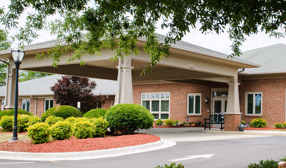 Covered porte-cochere and front entrance of a brick senior living building with manicured shrubs and a driveway.