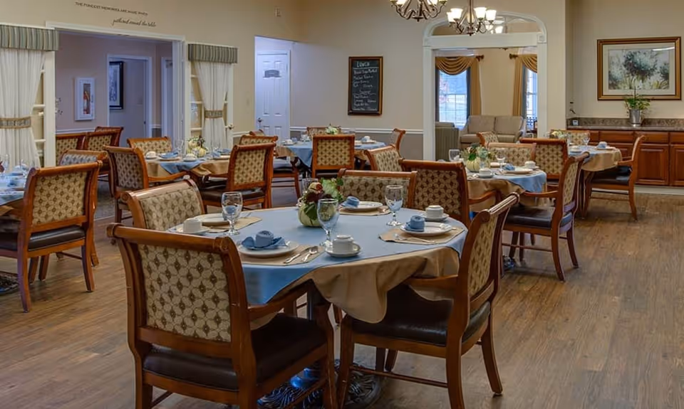 A dining room in a senior living facility with multiple round tables set with blue and beige tablecloths, white plates, cups, silverware, and glasses. The room has wooden chairs with patterned upholstery, wooden flooring, and soft lighting from chandeliers. There are decorative plants on the tables and framed artwork on the walls.