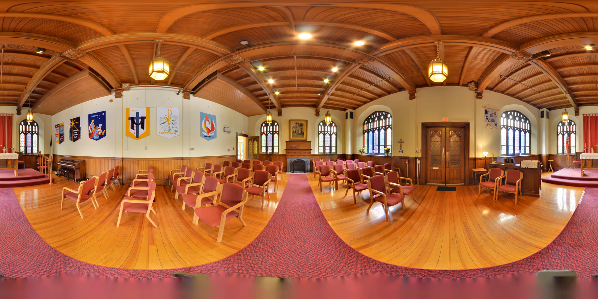 Interior panoramic view of a chapel or worship room with wooden ceiling beams, stained glass windows, wooden chairs with red cushions arranged in rows, banners hanging on the walls, and a red carpet running down the center aisle.