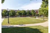 A large, well-maintained grassy courtyard area with a curved concrete walkway, surrounded by trees and lamp posts. In the background, there is a multi-story brick building with numerous windows under a partly cloudy sky.
