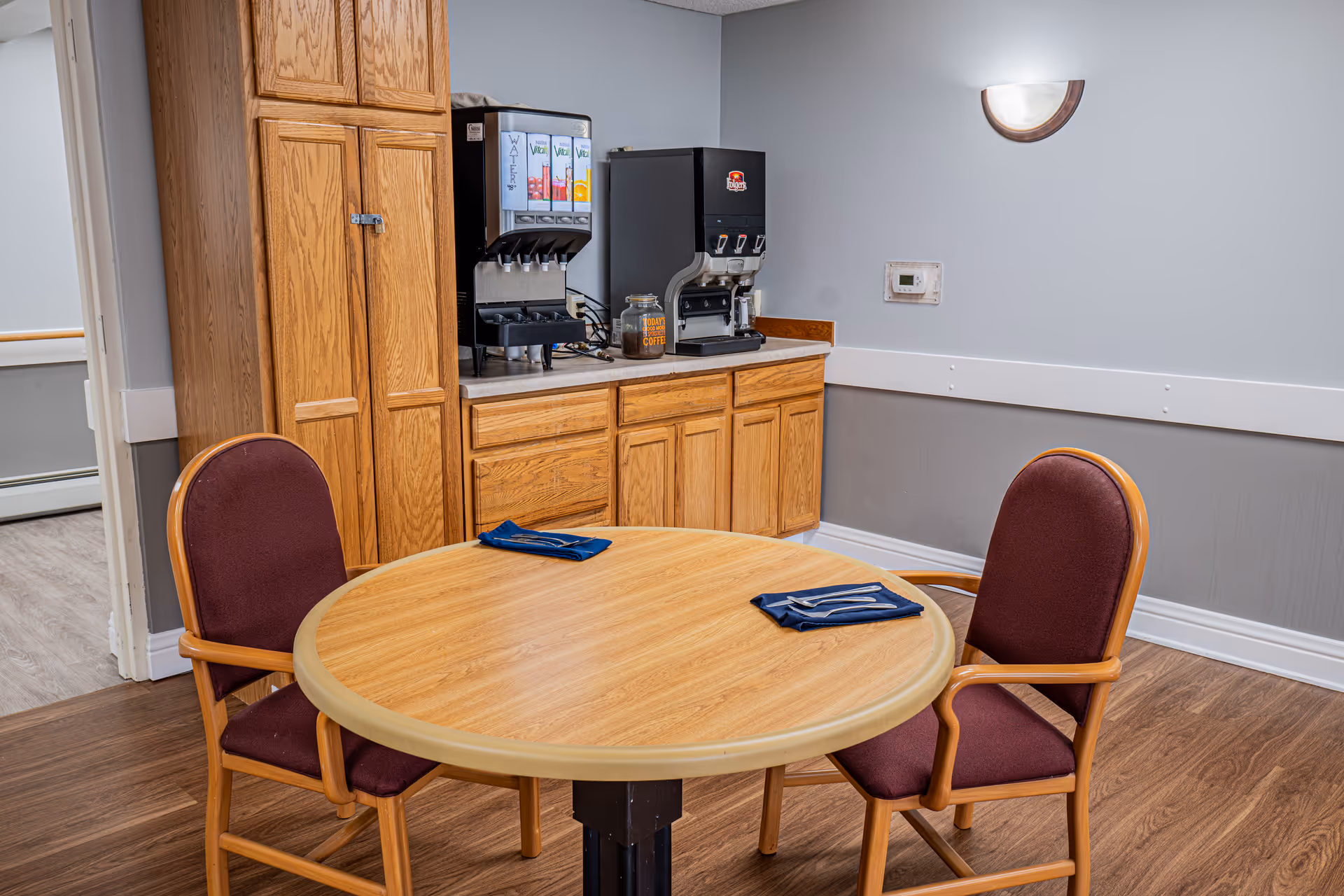 A small dining area with a round wooden table set with two folded blue napkins and two wooden chairs with maroon cushions. Behind the table is a wooden cabinet with a beverage dispenser and a coffee machine on the countertop. The walls are painted light gray with a white chair rail and a wall-mounted light fixture.