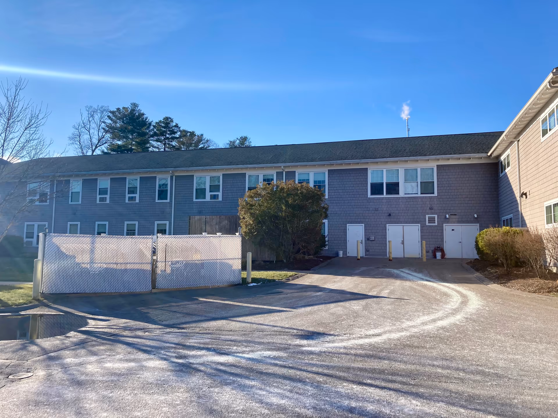 Exterior view of a two-story gray senior living building with a fenced area, driveway, and shrubbery under a clear blue sky.