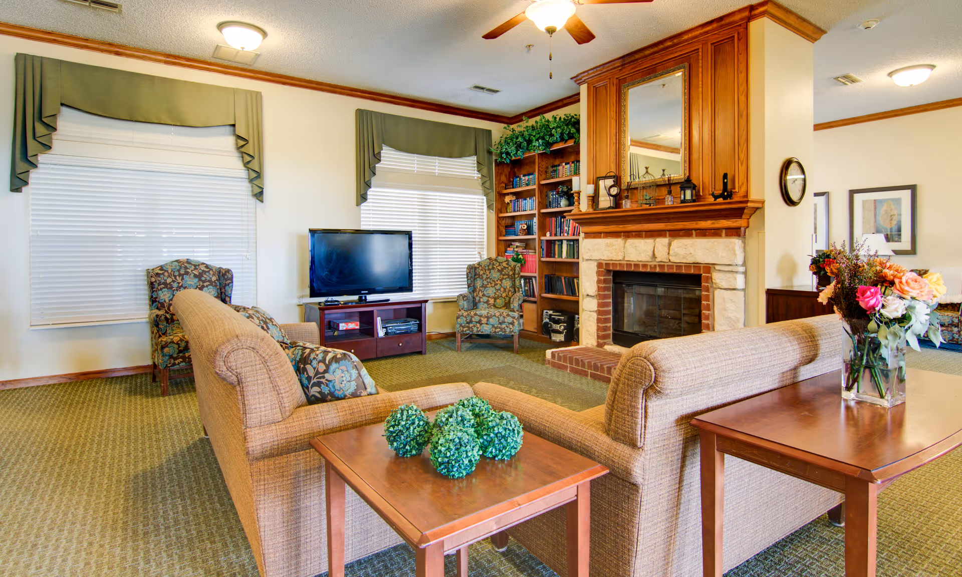 Cozy communal living room with two sofas facing a TV, armchairs, a fireplace with built-in bookshelves, and a side table with flowers.